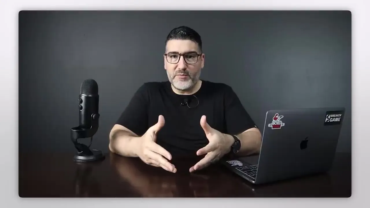 El profesor hablando a cámara en su mesa de trabajo con micrófono y portátil, explicando con las manos abiertas